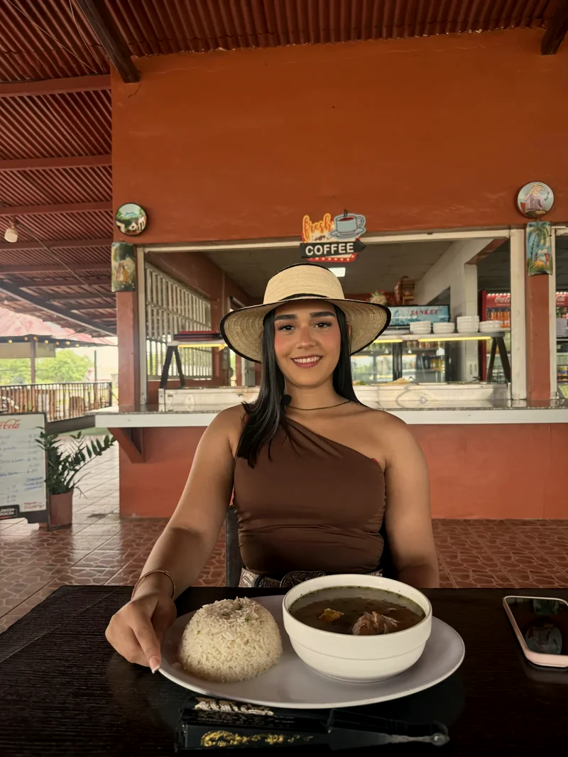 Mujer con sombrero de paja sentada a la mesa con sopa y arroz, frente a la barra de despacho