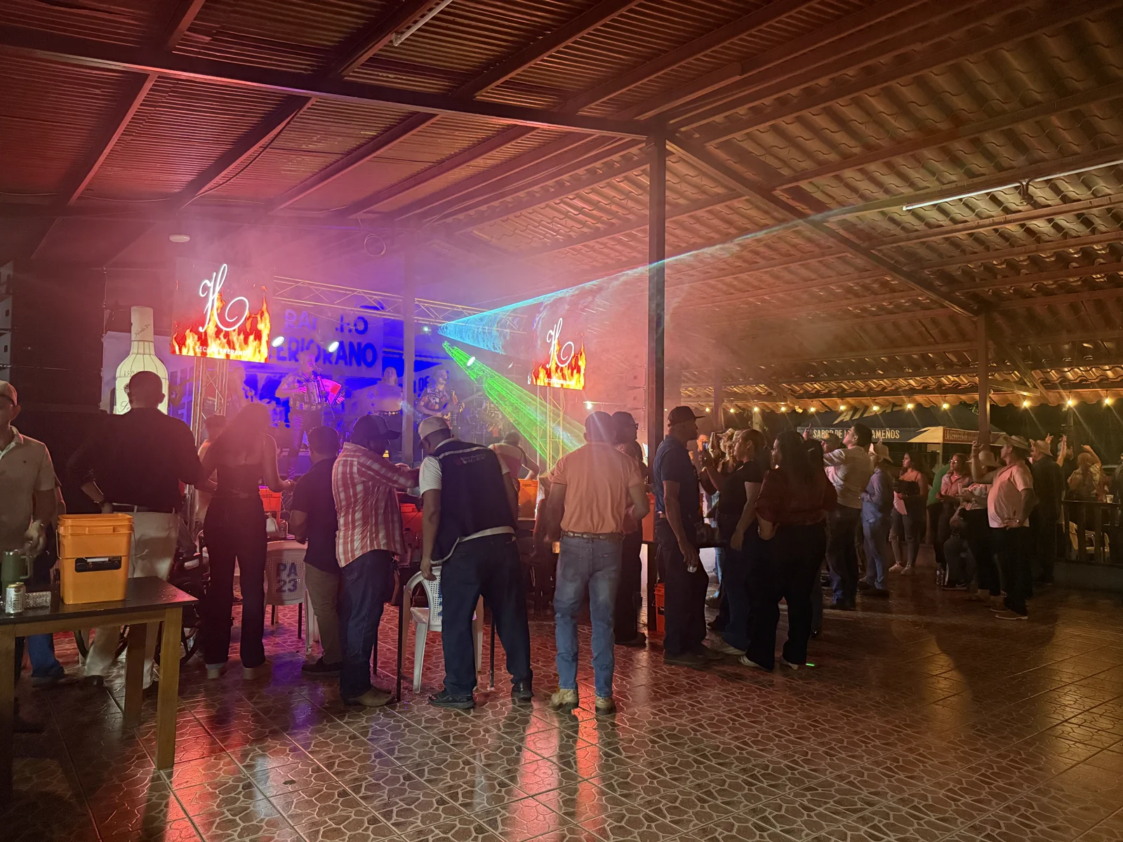 Vista amplia del rancho lleno de gente durante una noche especial, con la tarima, luces láser y mesas repletas