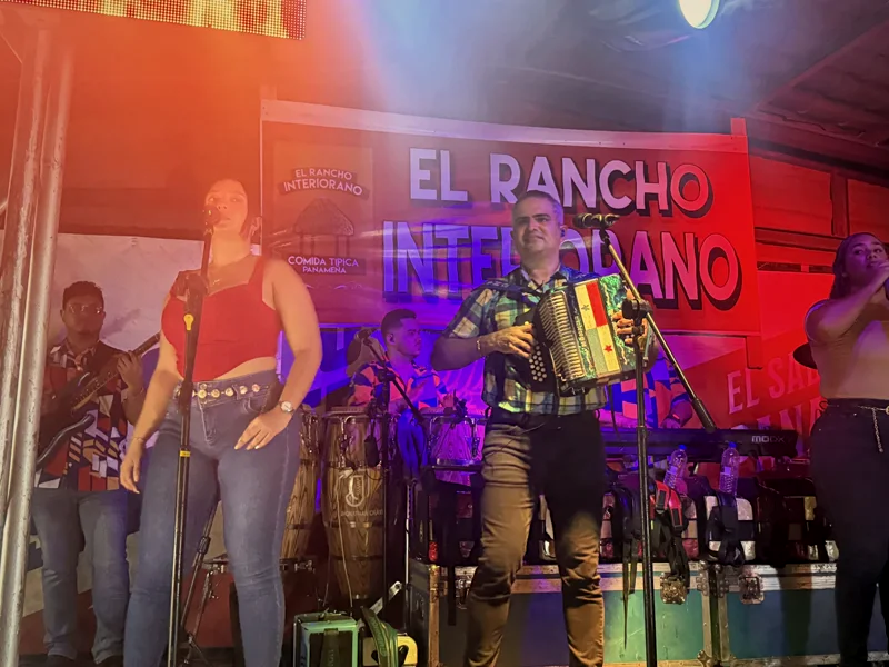 La orquesta completa tocando en la tarima frente al banner grande de El Rancho Interiorano, con luces de escenario